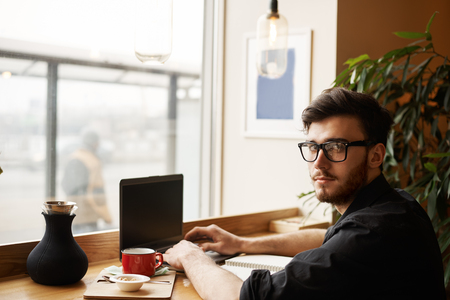 Freelancer working in cafe and drinking coffee or tea. Eye glasses man in black shirt sit in front of notebook and looking to the camera. hands on keybord typing text.の写真素材