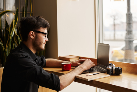 Young father in eye glasses search ideas of care little baby in the internet. Man visit cafe for a lunch time and surf with notebook and free wi-fi. Tourist write notes in his blog.の写真素材