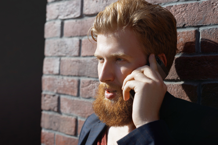 Red beard man talk by phone with happy face. Stylish caucasian dress hipster call friend and talk happy and good news. Sun effect. Close up phone conversation concept. Industrial part of the city.の写真素材