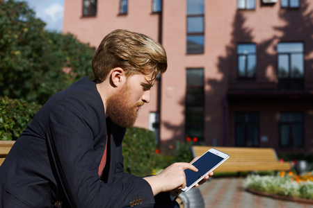 Handsome hipster use tablet for search a job. Man sit in industrial part of the city. Manager surf social network outdoor in break at work. Serious guy planning day in online schedulerの写真素材
