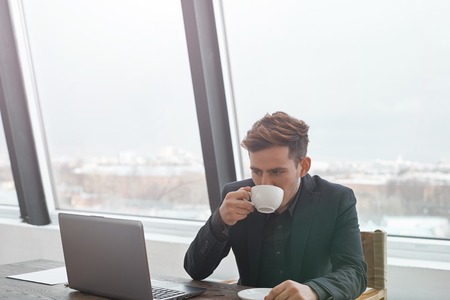Hardworking caucasian worker in stylish suite have time break for a cup of hot and tasty coffee. Relaxing on working place. Guy in office with glass wall and notebook and notepad on the wooden table.の写真素材