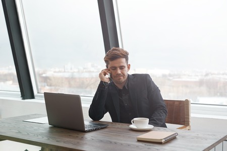 Young successful businessman in casual suit talk on his mobile phone while sitting in modern office, wealthy man having cell conversation with copy space area for content in glass windowsの写真素材