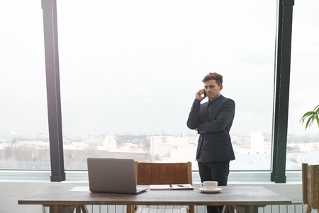 Portrait of successful businessman talking on mobile phone while standing against window in hallway of modern office interior, young confident man having cell telephone conversation during work breakの写真素材