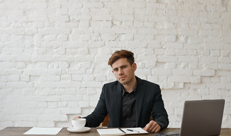 Good dressed manager sitting at the table with white cup of hot and tasty coffee near notebook and look to the camera. Brick wall backgound in loft office style. People and business concept.の写真素材
