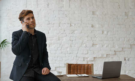 Young attractive engineer have serious phone call in the loft office. Man sitting on the wooden table with notebook and white brick wall background.の写真素材