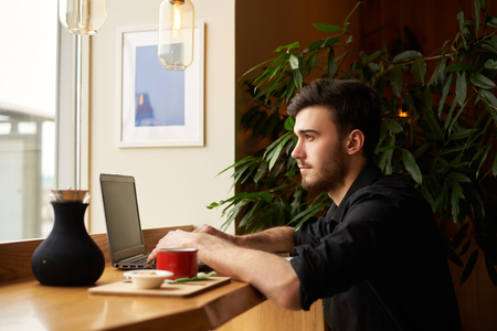 Young freelancer working in cafe with modern interior. Notebook connect to the internet through high speep wi-fi. Handsome guy look to the window on the street.の写真素材