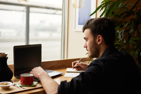 Checking mail or surfing social network concept. Man working online in beautiful place with cup of coffee. Modern guy in black tshirt sit under the table and typing on notebook new story of happy lifeの写真素材