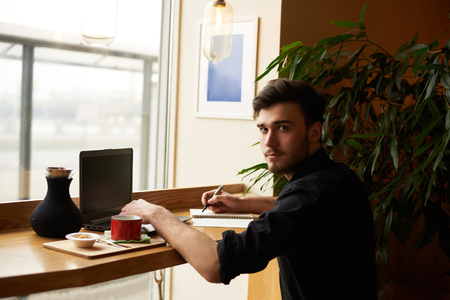 Modern dressed man sit in cafe with notebook and look to the camera with hands on laptop. Freelancer wait for the client for new job and use free wi-fi to surfing internet pages.の写真素材
