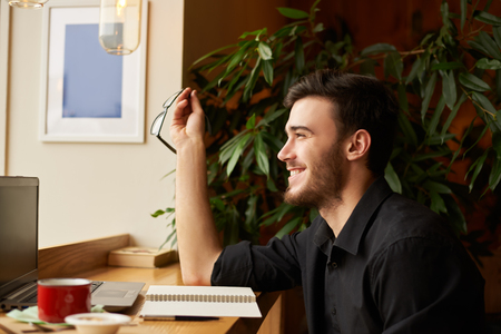 Side portrait of perfect look handsome caucasian man laughing and keep eye glasses in the hand. Guy get good news in the internet. Talented male editor start working morning in cafe.の写真素材