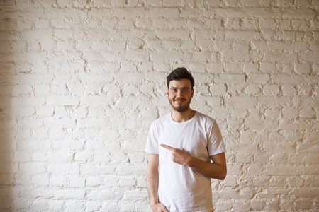 Casual dressed student point to the right side and stay in front of white brick wall. Portrait of handsome caucasian man look to the camera with loft interior. Copy space for advertising text.の写真素材