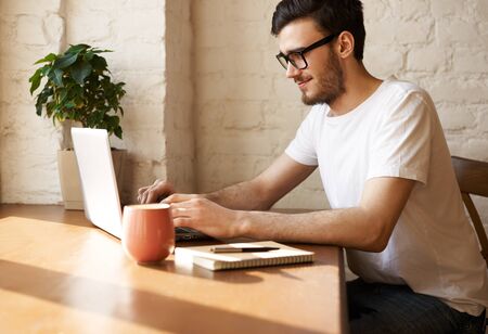 Young bearded journalist in eye glasses write new article in the internet and answer questions of his readers. Man in sunny day at home surfing social network and read news. Happy guy working indoor.の写真素材