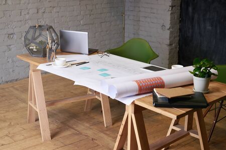 Architect workplace in the room. Business concept of architecture workshop. Constraction drawings on the table with notebook, phone. Loft interior with grey brick wall and blackboard.の写真素材