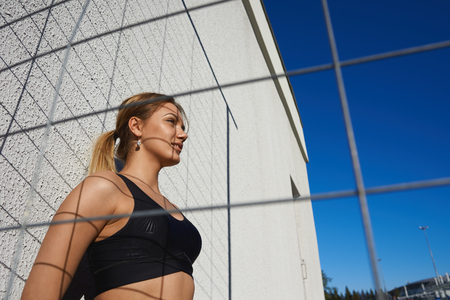 Low angle view of beautiful young Caucasian woman runner wearing black top relaxing after running workout in the morning, preparing for marathon, having determined look. Fitness, sports and trainingの写真素材