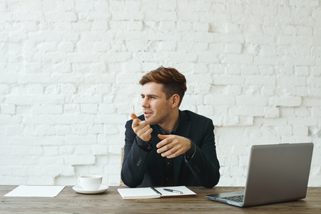 Attractive confident young recruiter sitting at office desk with open laptop computer, copybook and papers, looking sideways and pointing index finger at camera during job interview with applicantの写真素材