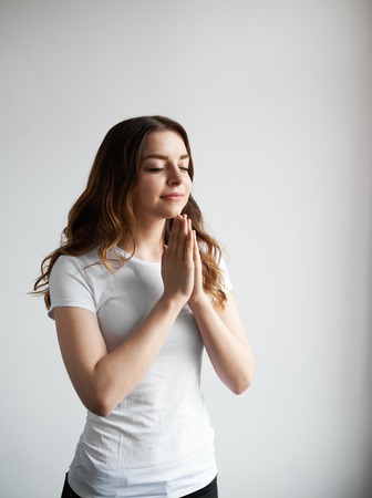 Positive girl wearing white casual t-shirt holding hands in namaste or prayer, smiling and keeping eyes closed while meditating and practising yoga indoors, having calm expression on her pretty faceの写真素材