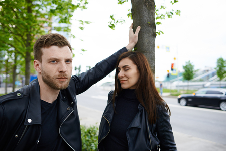 Beautiful young couple having walk in city park. Handsome stylish unshaven male in leather jacket standing close to pretty female, leaning hand on tree and looking at camera with serious expressionの写真素材