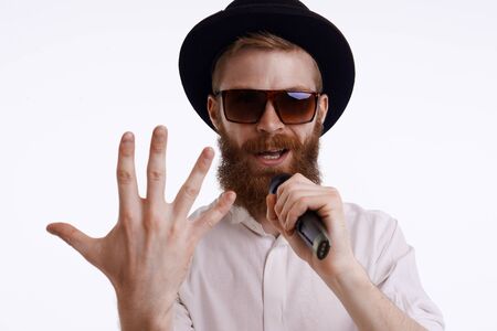 People, music, show and entertainment concept. Handsome stylish bearded showman or presenter wearing trendy hat and sunglasses performing on stage holding microphone, showing back of hand at cameraの写真素材