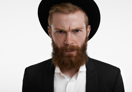 Headshot of grumpy young European hipster with fuzzy ginger beard frowning, having unhappy or angry expression on his face, feeling displeased and dissatisfied with something, wearing hat and suitの写真素材