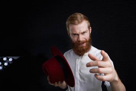 People, show and entertainment concept. Studio shot of macho man with fuzzy red beard and stylish haircut holding trendy hat and pointing finger at camera, presenting something or giving speechの写真素材