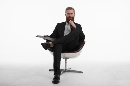 Live broadcasting. Portrait of confident young male professional news reporter with red beard sitting in chair in relaxed pose, holding clipboard and talking in microphone, dressed in stylish suitの写真素材