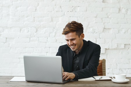 Stylish young European office worker in formal wear sitting in front of laptop pc, playing video games during lunch break, looking at screen with cheerful smile. Male employee working at officeの写真素材