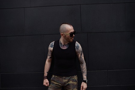 Confident serious young Caucasian bearded biker with mohawk and muscular tattooed arms posing...