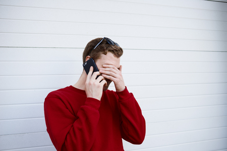 Bearded young Caucasian male in sweater having phone conversation with his friend, making facepalm gesture while laughing at funny story or joke. Modern technology and communication conceptの写真素材