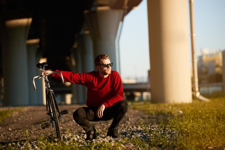 Handsome guy in trendy shades sitting in squatting posture on grass, holding his fixed gear bicycle, relaxing after long ride. People, leisure, hobby, active healthy lifestyle and transportationの写真素材