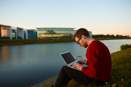 Outdoor shot of hardworking young European student in sunglasses studying remotely on notebook pc, using free city wi-fi, sitting on grass by the lake in public garden, having concentrated lookの写真素材