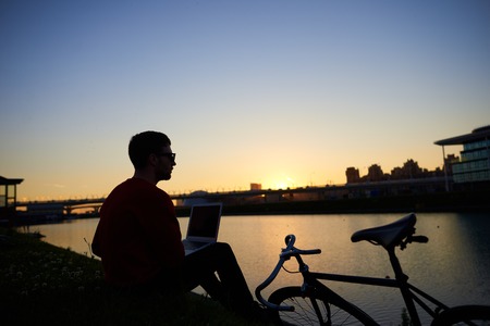 Silhoutte of hipster sitting on river bank with laptop pc on his lap, admiring amazing sunset after he finished work, feeling relaxed and connected to Universe. People, modern lifestyle and technologyの写真素材