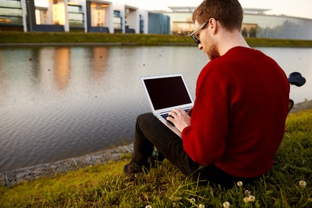 Rear view of stylish young male traveler sitting by the lake booking plane tickets online using free wi-fi on generic laptop computer with copy space blank screen for your advertising contentの写真素材