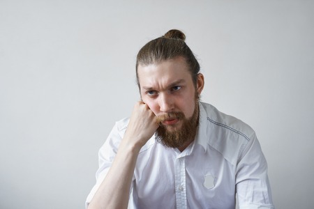 Sadness and boredom concept. Studio shot of unhappy young manager with hair knot and thick beard having upset and bored look, feeling tired and pissed off with boring working days at officeの写真素材