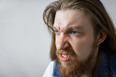 Close up studio portrait of angry mad young man with thick beard and long hairstyle frowning and clenching teeth, his eyes full of anger and fury. Negative human emotions, aggression, rage and frenzyの写真素材