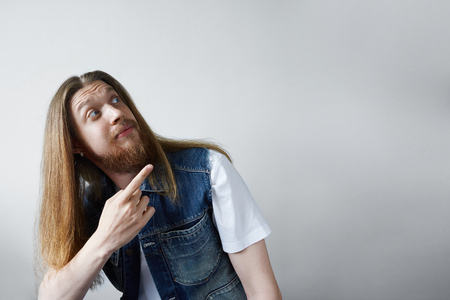 Indoor picture of funny unshaven young Caucasian man with long loose hair looking upwards and pointing index finger at grey blank studio wall with copy space for your text or promotional contentの写真素材