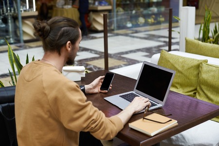 Modern stylish student wearing his long hair in knot using free wifi on his generic electronic gadgets with blank copy space screens for your content, surfing internet while waiting for friend at cafeの写真素材