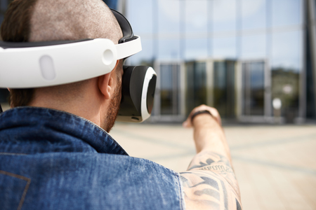 Rear view of brutal bearded European male hipster with tattoos and Mohawk hairstyle standing outdoors wearing futuristic 3d goggles with head mounted display and pointing finger at modern buildingの写真素材