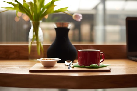 Mock up of hot coffee latte or cappuccino in red cup and white bowl with cane sugar cones resting on wooden table with decorative plants by the window glass in cozy restaurant during breakfastの写真素材