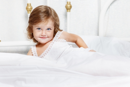 Sweet toddler lie in bed covered with white blanketの写真素材