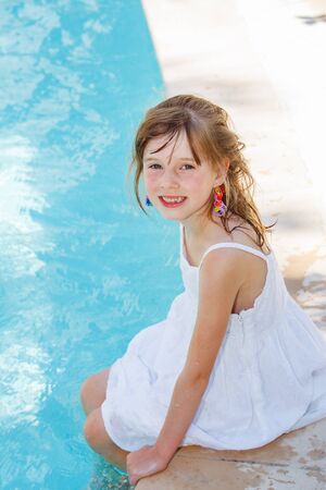 Smiling preschool girl in a white dress sitting by an outdoor swimming-pool, with her legs in waterの写真素材