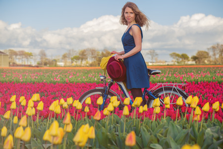 Happy female tourist in a blue dress standing with her bike in a spring field with yellow and pink  tulipsの写真素材