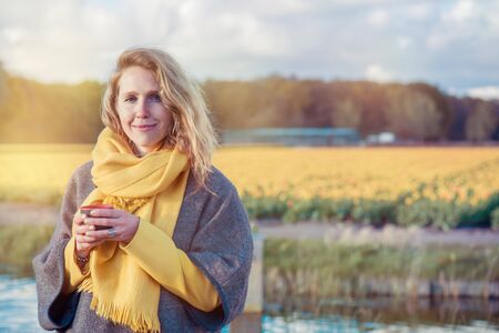 Portrait of a serene woman in  poncho and scarf holding a  cup with hot tea in the countryside. In the background  yellow tulip fields are situated.の写真素材