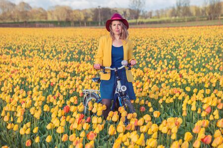 Smiling woman standing with her bike in a yellow and red tulips field at sunset.の写真素材