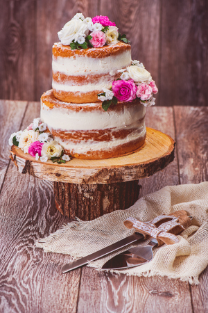 Homemade wedding naked cake decorated with flowers on wooden stand and serving set besideの写真素材