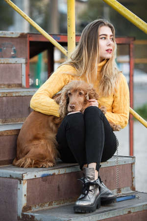 Portrait. A beautiful sexy girl, blonde, a young woman, looking like Jennifer Aniston sitting with a Cocker Spaniel dog, on a rusty metal stairwayの写真素材
