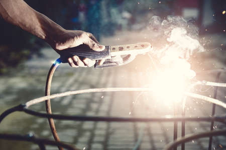 Young man worker welding iron pieces at work with protective goggles. Building construction concept.の写真素材
