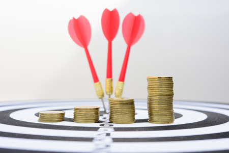 Red dart arrow hitting in the target center of dartboard with money gold coins.の写真素材