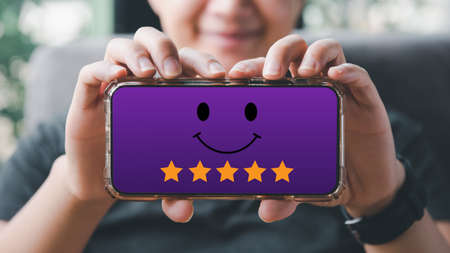 Customer satisfaction concept.Woman hand holding smartphone Show rating with five stars gold symbol and Smiley Face for her Satisfaction.の写真素材