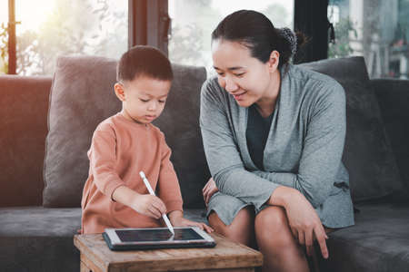 Home learning concepts. Asian mother is sitting next to her 3 year old son and she is teaching her children to learn via a tablet.の写真素材