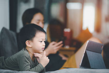 Asian boy looking at the tablet screen attentively. overstimulated children concept. Too much screen time. 3 years boy watching videos while tv is working. Internet addiction concept.の写真素材