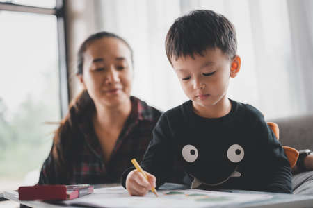 Asian Boy Doing homework with the intention and he mother Sitting beside. Child boy holding pencil writing. Distance Education concept.の写真素材